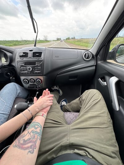 The image shows an interior view of a car, likely in motion, with a view of the road and fields through the windshield. The location is identified as Novomyshastovskaya, Russia. The windshield wipers are active, indicating rain. The dashboard and center console of the car are visible.

Two people are present. One person is wearing olive green pants and blue and white sneakers. The other is wearing blue jeans. Their hands are clasped together. The person wearing jeans has a tattoo of koi fish on their forearm, and is wearing a bracelet.