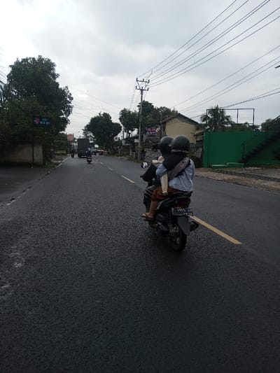 The scene depicts a street in the city of Kamji Na-Yekha, Bhutan, under an overcast sky. The dark asphalt road appears wet, possibly from recent rain, and features a faded yellow dashed center line. In the foreground, a dark-colored scooter with license plate F 4665 AD (06.24) travels on the right side of the road, partially over a broken yellow line fragment.
Three individuals are on the scooter. The driver, wearing a black helmet and dark attire, operates the vehicle. Directly in front of the driver, a small child wearing a black helmet faces forward. A rear adult passenger, also wearing a black helmet, a light blue denim-style jacket, brown trousers, and sandals, is seated behind the driver and carries a dark cross-body bag.
Further down the road, another scooter with a single rider (wearing a helmet) and a dark green truck are visible, both moving in the same direction as the foreground scooter.
The left side of the road is lined with trees and a blue rectangular sign displaying white text, possibly "KOPI" and "MINUM," along with a graphic resembling a coffee cup or face. On the right side of the road, behind the scooter, residential or commercial buildings are visible. One notable structure is a vibrant green-painted building with black exterior stairs leading to an upper level. Multiple utility poles with overhead power lines span across the upper portion of the frame.