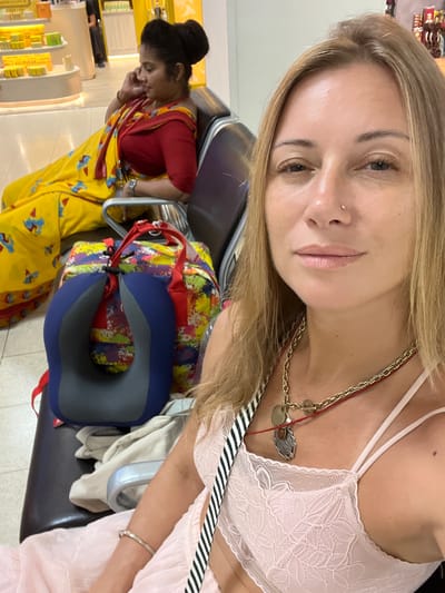 In Katunayake, Sri Lanka, two women are seated in a waiting area, likely at the airport, based on the presence of a neck pillow and luggage. In the foreground, a blonde woman is taking a selfie. She is wearing a pink lace camisole and a multi-layered necklace. Behind her, a woman dressed in a traditional yellow sari with a red blouse is seated, looking down and speaking on her phone. A colorful duffel bag and a blue neck pillow are positioned between the two women. The background shows shelves stocked with products, suggesting a retail environment.
