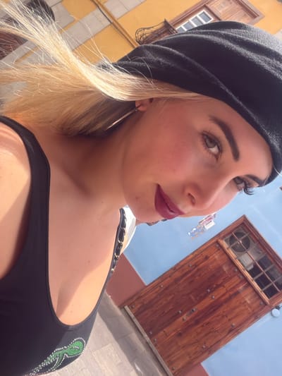 The image is a close-up, low-angle selfie of a woman with blonde hair, wearing a black beret and a black tank top. She is outdoors, with buildings in the background. One building is yellow with a white-framed window, and another is blue with a large, weathered wooden door featuring a small, barred window and a ventilation grille. The woman has dark lipstick and long eyelashes. Her blonde hair is blowing in the wind. The scene suggests a sunny day in San Cristóbal de La Laguna, Spain, given the architectural style and the possibility of vibrant building colors common in the Canary Islands.