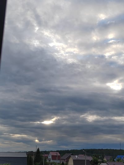 This is a landscape photograph taken outdoors. The sky is filled with a mixture of dark, heavy clouds and patches of bright sunlight breaking through. The clouds create a dramatic and somewhat overcast atmosphere. Below the sky, a village or rural settlement is visible. Several houses with different roof colors, including grey, red, and brown, are spread across the landscape. Some houses have visible chimneys. Trees, including evergreens and deciduous trees, are scattered throughout the village and form a dense forest in the background. There are also visible structures that appear to be utility poles or small towers. The overall scene suggests a daytime setting, likely late afternoon or early evening given the cloud cover and the quality of light. The weather appears to be changeable, with the sun intermittently shining through the clouds. The location is identified as Zhal'skoe, Russia. There is no discernible text in the image. The image evokes a sense of a quiet, rural setting under a dramatic sky.