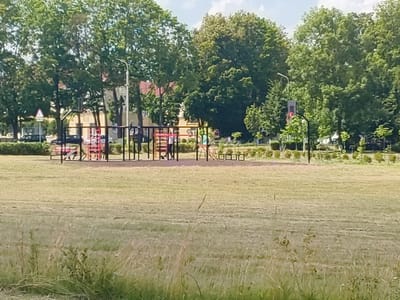 This image depicts a playground scene in Klichaw, Belarus. The playground features red and silver metal structures, including a climbing frame and swings. In the background, several people are visible on the playground, appearing to be engaged in recreational activities. The setting is a park-like area with a large, mowed lawn in the foreground and a line of mature trees behind the playground. Beyond the trees, buildings and a street with parked cars are visible, suggesting an urban or suburban environment. The sky is partly cloudy, indicating fair weather. The time of day appears to be daylight. There is no discernible text in the image.