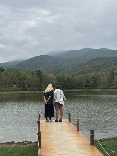 Two individuals stand on a wooden pier extending into a body of water. One person, closer to the edge of the pier, has long blonde hair and wears a black top and skirt. The other person, standing slightly behind, wears a white top and dark shorts. Mountains covered with green vegetation rise in the background under an overcast sky. The scene is located in Ban Huai Sai, Thailand.