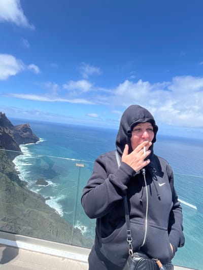 A medium shot, taken in vertical orientation, captures a person in a black Nike hooded sweatshirt smoking a cigarette. The person is positioned on a glass-walled overlook, offering a clear view of the azure ocean and coastline below. The sky is a clear blue, dotted with fluffy white clouds. The location is in the city of La Aldea de San Nicolás, Spain.