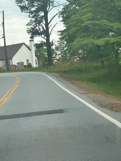 The image shows a view from a vehicle traveling on a road in Mountain Park, United States of America. The road has double yellow lines in the center and a white line on the right edge. To the left of the road, there's a grassy area with trees and a house with a white exterior and a brown roof. Utility poles and wires are also visible. The sky is overcast.