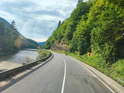 This media file captures a scenic view of a winding road alongside a river, flanked by lush green forests on a mountainside. The road, paved and marked with a white center line, curves to the right, following the path of the river. A metal guardrail separates the road from the riverbank, which is lined with rocks and vegetation. The river itself appears to be flowing briskly, with some whitewater visible.

On the right side of the road, a steep, rocky embankment rises, covered in dense green trees. Two yellow and black directional signs, indicating sharp curves to the left, are visible along the roadside. The sky above is partly cloudy, with sunlight breaking through, suggesting it is daytime, likely late morning or early afternoon. The overall atmosphere is one of natural beauty and tranquility, with a sense of journey implied by the road. The location is identified as Andreneasa, Romania. There are no people or specific activities depicted, focusing solely on the natural landscape and the road.