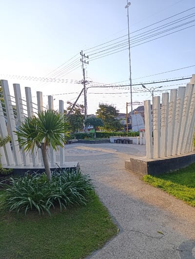 The scene is a public park in Kecamatan Asembagus, Indonesia, located at coordinates -7.75 latitude and 114.218 longitude. The park has a paved walkway that leads from the viewer's vantage point to a circular area in the center of the park. There are white vertical slats on either side of the walkway. To the left, there is a large palm tree and a patch of green grass, as well as several smaller plants. To the right of the walkway, there are more green plants and trees in the distance. In the background, there are power lines and a tall utility pole with a radio antenna on top. The sky is blue and clear, with the sun shining. There are no people or animals visible in the scene.