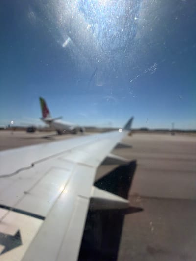 The image shows a view from an airplane window, looking out at the wing and the sky. The wing is white and extends diagonally across the frame. The sky is a deep blue, with some light reflections on the window. In the distance, another airplane's tail can be seen, with green, red, and white markings. The ground below the wing is a light grey tarmac. The sun appears to be bright, casting a glare on the window, suggesting it is daytime. The scene appears to be at an airport or during flight, with the context suggesting Moreira, Portugal. There is no visible text in the image. The overall impression is one of travel and observation from an aerial perspective.