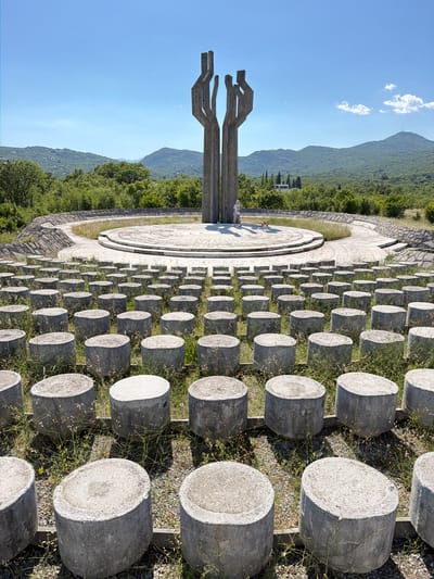 The image depicts the Barutana Memorial Complex in Montenegro. It features a large, circular concrete plaza and a series of concrete cylinders arranged in rows. A concrete abstract sculpture stands adjacent to the plaza. The landscape includes grassy areas, trees, and distant mountains under a blue sky. A person is present on the plaza.