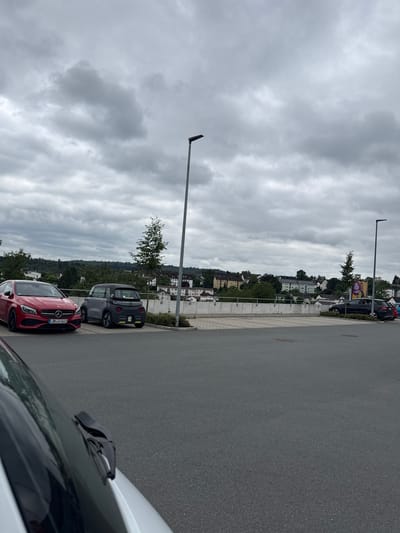 The image shows a parking lot in Braunfels, Germany, under a cloudy sky. In the foreground, a portion of a white vehicle's windshield and wiper blades is visible, angled towards the right. To the left of the image, a red Mercedes-Benz CLA-Class vehicle is parked, displaying the license plate "LDK S 4621". Next to it, a small, dark gray electric car with yellow accents is parked. In the background, a line of trees and buildings forms a cityscape on a hillside. A lamppost stands to the right of the cars, and another lamppost is visible further in the background near a billboard. The terrain is paved with asphalt.