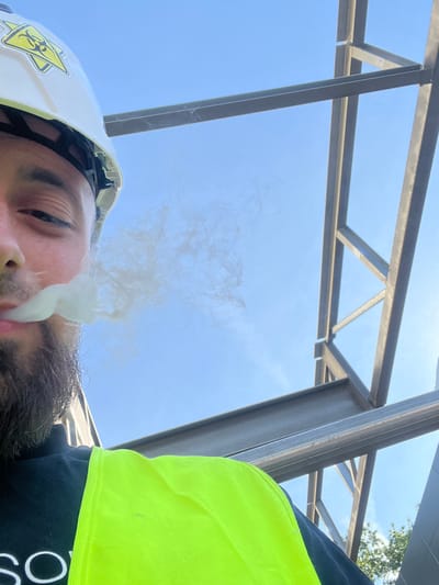 A close-up, low-angle shot features a person wearing a white hard hat with a yellow biohazard sticker and a neon yellow safety vest over a dark shirt. The person is exhaling a cloud of white vapor. The background consists of a clear blue sky and the steel framework of an unfinished building, suggesting a construction site. The image appears to be taken outdoors. The location is identified as Jabłonna, Poland.