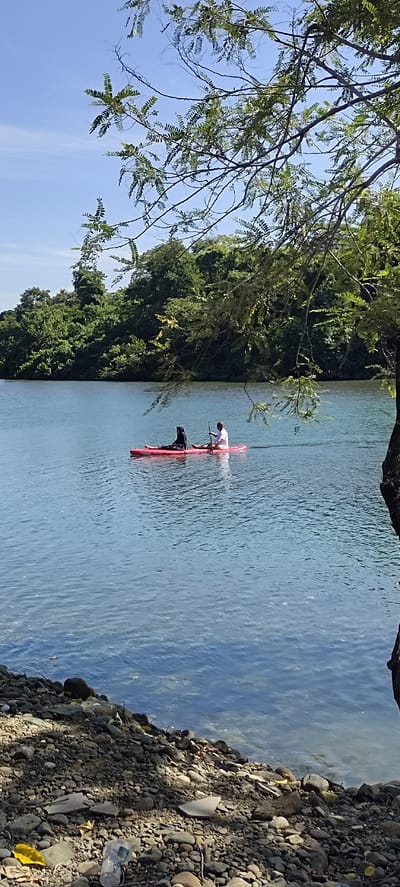 Two individuals are paddleboarding on a tranquil, clear blue river. One person, dressed in a black hijab and attire, is seated towards the front of the red paddleboard, while the other, in a white shirt and shorts, is seated towards the back, holding a paddle. The river reflects the bright blue sky and is bordered by lush green trees on the opposite bank. The foreground features a rocky shoreline with scattered debris, including a yellow leaf and a clear plastic cup. A tree with green leaves partially obscures the view in the upper right corner of the image. The scene suggests a serene natural setting, possibly in the city of Lamseunia, Indonesia.