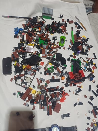 The image shows a pile of Lego building blocks and other small toys scattered on a white surface, possibly a bedsheet. Among the Lego pieces, there is a green toy car, a black rectangular object, a pencil with "HIGH QUALITY PENCIL" written on it, and a silver pen. The objects are spread out and appear to be in disarray, suggesting they have been played with. There are no people visible in the image. The lighting is bright, casting shadows on the white surface. There is no information to suggest this scene is located in Jumashuy, Uzbekistan.