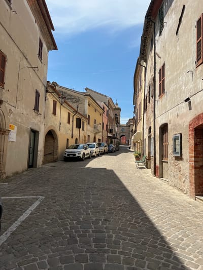 A cobblestone street runs between two rows of aged buildings in Barbara, Italy. The buildings on the left are primarily beige, with arched doorways and wooden shutters on the windows. On the right, the buildings are a mix of beige and brick, also featuring shutters and visible architectural details like exposed brickwork and arched entrances. Several cars are parked along the street, and a lone figure walks in the distance towards an archway that frames a clock tower. The sky above is clear and blue, casting distinct shadows across the pavement. A green bench sits outside one of the buildings on the right.