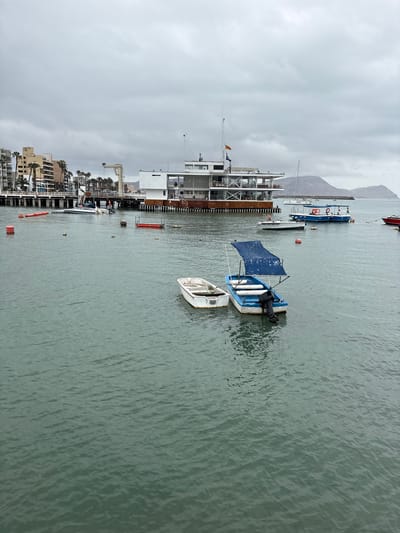 The image shows a cloudy day at a harbor in La Punta, Peru. In the foreground, two small boats are floating on the water. The boat on the left is white and appears to be unoccupied. The boat on the right is blue and white, with a blue canopy and an outboard motor. It also appears to be unoccupied.
In the midground, a larger white building with multiple levels and large windows is situated on a pier. A white sailboat is docked near this building. Several smaller boats and buoys are visible in the water around the pier.
In the background, several multi-story buildings with palm trees in front of them line the shore. Further in the distance, a mountain range can be seen under the overcast sky. Seagulls are flying in the sky. The water appears calm with gentle ripples.