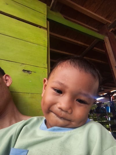This is a selfie taken by a young child in Pompanua, Indonesia. The child is wearing a light green t-shirt with a blue collar. Behind the child, a wall of weathered green wooden planks is visible. The roof structure above appears to be made of wood. In the background, shelves stocked with various items can be seen, suggesting an indoor or semi-outdoor storage area. The overall lighting suggests it is daytime.
