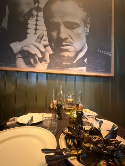 The image displays an indoor dining scene within a restaurant located in Lillestrøm, Norway. A dark wooden table in the foreground holds the remnants and setup for a meal. On the left, a large empty white oval platter and a smaller, mostly cleared white plate with a knife are visible, alongside a clear glass tumbler containing water. Centrally, a white rectangular dish is filled with cooked mussels in their shells, some submerged in a yellowish broth, with lemon pieces and serving utensils. Further on the table, a stemmed glass holds white wine, with another similar glass partially obscured. Two amber-colored bottles, consistent with beer bottles, and an empty clear glass bottle are present. A decorative gold-colored lantern with intricate cutouts provides ambient light, flanked by two dark-colored salt and pepper grinders. Above the dining area, a large framed black and white portrait of Marlon Brando as Vito Corleone from *The Godfather* is mounted on the wall. The lower section of the wall features dark green vertical wooden paneling. No individuals are visible in the scene.