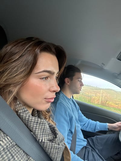A photograph taken from inside a vehicle depicts two individuals, a female and a male, traveling in Mafra, Portugal. The female, in the foreground, occupies the front passenger seat, secured by a visible seatbelt. She has light brown, wavy hair and is seen in profile, looking towards the right. She wears a grey and white checkered scarf and a brown outer garment. Behind her, the male occupies the driver's seat, also secured by a seatbelt. He has dark hair and wears a light blue long-sleeved shirt and dark trousers; his right hand is visible on the steering wheel, and he is looking forward. Through the vehicle's windows, an exterior rural landscape is visible, featuring rolling hills with green and brown vegetation, indicative of fields or agricultural land, and utility poles in the mid-distance under a bright, possibly overcast sky.