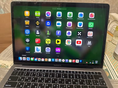 The image shows a MacBook Air laptop displaying a screen full of application icons. The laptop is open and resting on a patterned surface. The screen displays a green background with a search bar at the top. Below the search bar are rows of app icons, including Duolingo, Microsoft Word, Microsoft Excel, WhatsApp, and others. At the bottom of the screen, there is a dock with more application icons and a trash can. The keyboard of the MacBook Air is visible in the foreground. The overall scene suggests a casual setting, possibly a home or office environment. There are no people visible in the image. The location of Демихово, Russia is not visually identifiable from the image content.