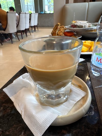 The image shows a close-up view of a glass of coffee on a saucer with a white napkin, placed on a dark, reflective surface. In the background, several white chairs with wooden legs are visible, some of which are occupied by people. A bottle of water is partially visible to the right of the coffee glass. Further in the background, there are glimpses of tables with food and drinks, suggesting a dining or cafe setting. The environment appears to be indoors, with natural light filtering in from large windows. There is no specific indication of the location being Lengkong Kulon, Indonesia, based on the visual information provided.