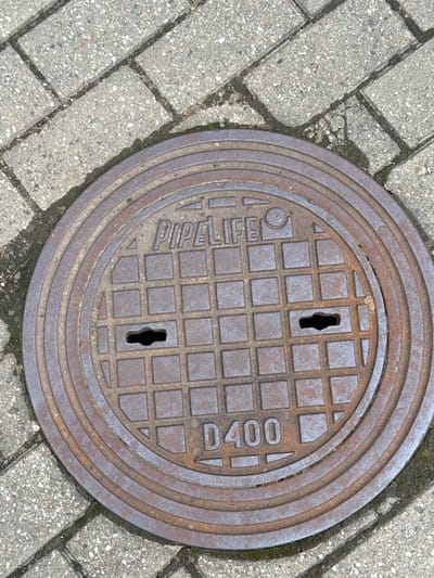 An overhead view shows a circular, rusted metal manhole cover embedded in a paved surface. The manhole cover has concentric rings and a grid pattern. In raised lettering, the words "PIPELIFE" are visible across the top of the cover, and "D400" is visible across the bottom. There are two keyhole-shaped openings in the grid pattern. The surrounding paving stones are gray and rectangular, laid in a diagonal pattern, with dark grout lines and some sparse green moss. The scene is well-lit, with no people or animals present. The location is Upeslejas, Latvia.
