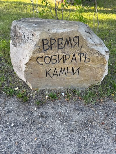 A large, rough-hewn stone, likely a boulder, rests on the ground in a patch of grass and dirt. The stone has been carved with Russian text. The inscription reads "ВРЕМЯ СОБИРАТЬ КАМНИ" which translates to "It is time to gather stones." The stone is positioned outdoors, with a background of green grass and some sparse vegetation. The ground in front of the stone is a mixture of asphalt and gravel, with scattered dry leaves and twigs. The lighting suggests it is daytime, possibly late afternoon due to the warm light. There are no people or animals visible in the image.