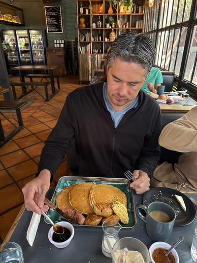 Here is a description of the image:
A man is sitting at a table inside a restaurant, preparing to eat a meal. The man has graying hair and is wearing a black jacket over a blue shirt. He is using a fork and knife to cut into a stack of pancakes, which are served on a metal tray with other breakfast items, including sausage and scrambled eggs. There is a small dish of syrup next to the pancakes. On the table, there is also a mug of coffee, a plate with remnants of food, a small bowl of sauce, and a glass with ice. Another person wearing a tan jacket is seated at the same table.
The restaurant appears to have a rustic decor, with exposed brick floors and a wooden bookshelf filled with various items. A menu board and a refrigerated display case are visible in the background. The scene suggests a casual dining environment.
Based on the request for the location, the restaurant may be located in La Tijera, Mexico.