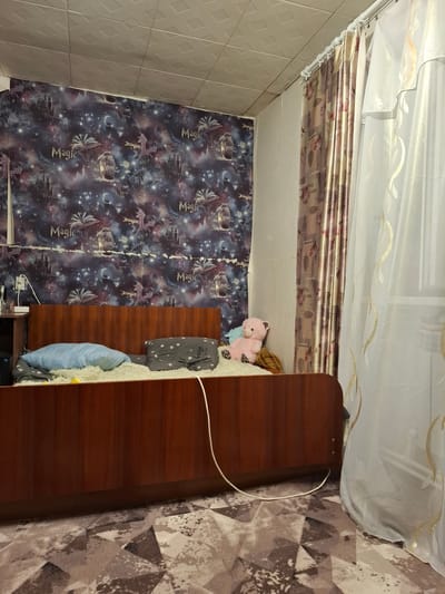 The image depicts a bedroom in Novlyanka, Russia. The room features a wooden bed with a headboard, covered with a fluffy white rug. Two pillows are on the bed: one light blue and another dark gray with a star pattern. A pink teddy bear sits on the bed near the pillows. The walls are decorated with wallpaper featuring a space-themed design with castles, ships, and the words "Magic" and "Imagine." To the right of the bed, there are curtains; one is patterned with floral motifs in shades of brown and gold, and the other is a sheer white curtain with gold leaf-like embellishments. The ceiling is paneled with a diamond pattern. A small wooden nightstand is visible to the left of the bed, with a dark object on it, possibly a computer tower, and a white electrical cord running from it. The floor is covered with a patterned rug in shades of beige, brown, and gray.