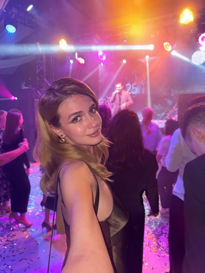 The image captures an indoor evening event or party in Редант, Russia. In the foreground, a young adult female with light brown hair, wearing a dark, possibly bronze, spaghetti-strap dress and a gold-colored earring, is taking a selfie with her right arm extended, looking towards the camera with a slight smile. The background reveals a brightly lit stage where a male performer in a light-colored suit is singing into a microphone. A large digital display showing the number '25' is visible behind the performer. The stage and event space are illuminated by multiple colorful stage lights, casting beams of pink, purple, orange, and blue light. Several other event attendees are visible, some partially obscured, standing and observing the stage. The floor is covered with numerous scattered metallic confetti pieces, reflecting the ambient light. White draped fabric is discernible near the ceiling as part of the event decor.