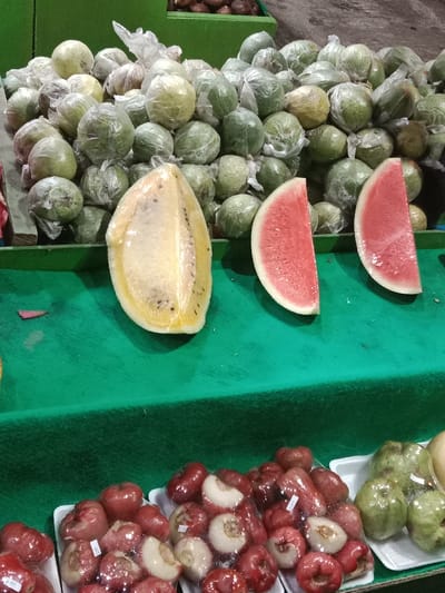 The image displays a produce stall in Sekopek Wetan, Indonesia. The upper portion of the frame features a large mound of light green, ovoid fruits, each individually encased in clear plastic film, piled on a surface behind a green structure. To the upper right, brown, textured fruits, consistent with Salak, are partially visible in a green container. The mid-ground reveals a green fabric-draped counter where three cut fruit sections are arranged: a large, pale yellow fruit slice with visible dark seeds, cut lengthwise, is positioned to the left of two smaller, red watermelon slices with white rinds, each cut into a quarter-circle shape. The lower foreground presents multiple white styrofoam trays. These trays contain red, bell-shaped fruits, identified as likely water apples or rose apples; some are whole, while others are sliced or peeled to expose lighter interiors. Each fruit in these trays is individually sealed in clear plastic film and bears a small white label. An additional white styrofoam tray to the right holds more light green, plastic-wrapped fruits similar to those in the upper pile. No individuals are present in the scene.