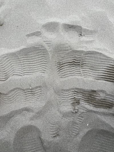 The image displays a close-up, top-down view of fine-grained, light-gray sand, consistent with a sandy beach in Ulcinj, Montenegro. The sand surface is marked by multiple impressions. Several distinct shoe or boot prints are visible, characterized by a ribbed tread pattern consisting of parallel, horizontal ridges, approximately 1-2 cm in width, often with a central longitudinal depression. These prints indicate foot traffic across the sand. A narrower, linear track, approximately 10-15 cm wide, runs generally vertically through the center of the frame, showing a segmented pattern consistent with a small tire or roller. This track appears to intersect and overlay some of the shoe prints. Discarded objects embedded in the sand include a light-tan cigarette butt located in the lower-left quadrant and a small, irregular white fragment, possibly a shell, in the lower-right quadrant. No individuals or other environmental features are visible.