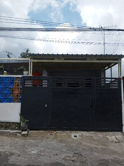 The image depicts the exterior of a residential building in the city of Viengthong, Laos, viewed from a slightly low angle on an overcast day with patches of blue sky. The central feature is a closed, dark grey or black metal gate comprising three sections: a wider double-door segment in the middle and a narrower single-door segment to its right. The gate features horizontal paneling in its lower half and horizontal slats in the upper half.

Above the gate, a carport or porch structure with a light yellow/gold metal frame supports a corrugated metal roof. To the immediate left of the gate, a wall section is clad with decorative hollow concrete blocks in alternating blue and brown/orange colors. Several potted plants are visible on ledges above this wall and peeking over the gate. Barbed wire is present on top of the left-most wall section. Behind the gate, a reddish object, possibly a storage tank, is partially visible.

The ground directly in front of the gate is an uneven, patched concrete surface. A small plant in a repurposed plastic bottle rests at the base of the left wall. A square drainage grate is embedded in the concrete near the center of the gate's base, and a small, light-colored rectangular object, possibly paper currency, lies on the ground to the left of the grate.

Above the building, numerous parallel black electrical or utility wires traverse horizontally across the frame against the sky. A television antenna is visible on the right side, extending above the roofline. No people, vehicles, or animals are present in the scene.