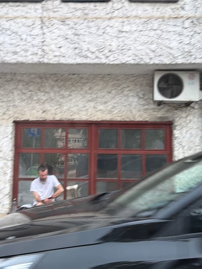 The image shows a close-up of a building's exterior, featuring a textured, light-colored wall. An outdoor air conditioning unit is mounted on the wall to the left. To the right of the AC unit is a wooden-framed window with multiple panes. Reflections are visible in the window glass.
A person, wearing a white t-shirt, is leaning out of the window. They have long brown hair and appear to be holding a phone or similar object. The person's face is partially obscured and their expression is not clearly discernible. The angle of the shot suggests the photo was taken from a car, as the shiny surface of a vehicle is prominently visible in the foreground, partially obscuring the view of the building and window.
The scene appears to be outdoors, with natural light suggesting it is daytime. The weather is not clearly indicated. There are no specific location cues beyond the provided context of Konjic, Bosnia and Herzegovina. The overall mood is casual and everyday. No visible text is present in the image.