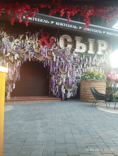 The image captures an outdoor establishment decorated with abundant floral arrangements and signage, potentially a cafe or bar. Dominating the facade are large, illuminated, white marquee letters spelling "СЫР," which translates to "cheese." To the left, an archway, also adorned with faux wisteria and small woven baskets, frames a dark, corrugated metal shutter, suggesting an entrance or window. Above the archway, a black banner displays white text, "КОКТЕБЕЛЬ," meaning "cocktail," repeated twice with a small bird graphic in between. Red artificial flowers hang from above, creating a canopy effect. To the right, a large bale of hay serves as a planter, filled with yellow and white flowers, and a black wicker chair is positioned nearby. Further to the right, a large red mushroom sculpture is visible. The ground is paved with grey rectangular tiles. The timestamp "19.06.2026 05:18" is present in the lower right corner. The location is identified as Kiparisnoe, Ukraine.