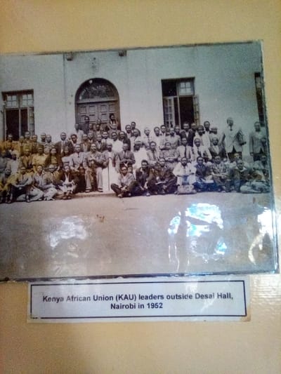 A framed, black-and-white historical photograph is affixed to a plain, light-colored wall. The photograph is covered by a reflective surface, causing a significant glare in the bottom right quadrant. Below the photograph, a white rectangular caption reads, "Kenya African Union (KAU) leaders outside Desai Hall, Nairobi in 1952."
The photograph depicts a large group of approximately 60 to 70 adult African men posing for a formal portrait outdoors. The subjects are arranged in multiple rows: a back row stands, middle rows are seated on chairs, and a front row is seated directly on the ground. The men are predominantly dressed in Western-style formal attire from the mid-20th century, including suits, collared shirts, and ties, with some individuals wearing simpler shirts or traditional garments. They are all looking towards the camera. The background consists of a light-colored, multi-story building, identified as Desai Hall. The building features a prominent central arched doorway with double wooden doors, flanked on either side by rectangular windows with multiple panes. The ground in the foreground appears to be a flat, unpaved surface. The event depicted in the photograph took place in Nairobi, Kenya. This physical image is located in the city of South Sakwa ward, Kenya.