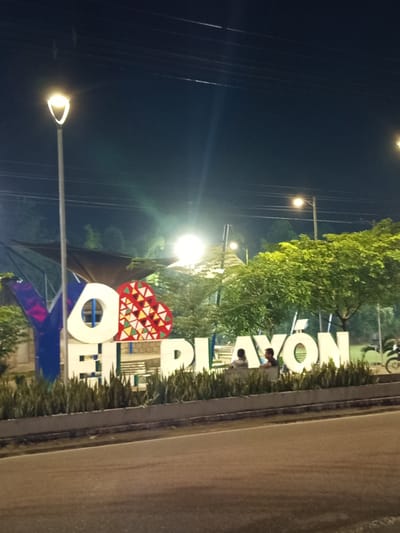 This is a nighttime, eye-level, medium shot of the "YO EL PLAYÓN" sign in El Playón, Santander, Colombia. The sign is illuminated by streetlights and is positioned in front of lush green trees. The large, white letters spelling out "EL PLAYÓN" are prominent in the foreground, with a stylized red, white, and yellow heart icon integrated into the design. The letters "YO" are partially visible on the left side of the frame, with the "Y" being blue and the "O" being white and circular. Two individuals, a male and a female, are seated on a bench behind the sign, facing towards the right. A motorcycle is parked to the far right of the frame. The background consists of dark trees and the night sky with visible power lines and streetlights. The scene appears to be a public park or recreational area within the city.