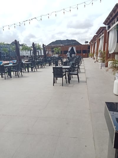 The image shows an outdoor patio area with black tables and chairs arranged in rows. There are white curtains hanging from the building to the right. The floor is tiled and the area is lit by string lights. There is a white, cylindrical object in the background, possibly a water tank. The image is taken from a low angle, looking towards the back of the patio. The location is a restaurant or bar in Ujevwu, Nigeria, specifically at the coordinates latitude: 5.472, longitude: 5.817. There are no people visible in the image.