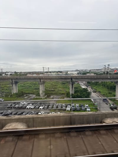 The image captures a view from a moving train in Gongshu District, China, looking out over a landscape dominated by an elevated train track and a concrete overpass. In the foreground, the tracks of the train from which the photo is taken are visible. Below the overpass, a large, open-air parking lot is filled with numerous cars, predominantly dark in color. Beyond the parking lot, a road runs alongside a densely vegetated area, with more cars and trucks on the road, including a large blue truck. The middle ground features the supporting pillars and deck of the overpass, with green fields and some scattered structures visible underneath and behind it. In the background, a city skyline with numerous buildings stretches across the horizon, under a cloudy, overcast sky. The scene suggests an urban or suburban environment with significant infrastructure for transportation and industry.