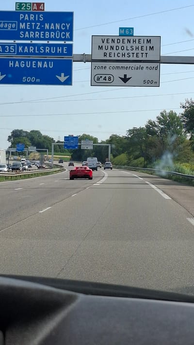 The image is a first-person perspective from a vehicle driving on a multi-lane highway. The view is directed forward and slightly downward. In the foreground, the dashboard of the vehicle is visible, colored black. The highway itself is paved with asphalt and has white lane markings.
On the left side of the highway, several blue road signs are mounted on poles. The topmost sign indicates "E 25 A 4" in green and white. Below it, a blue sign lists destinations: "PARIS," "METZ-NANCY," and "SARREBRÜCK." Further down, another blue sign reads "A 35 KARLSRUHE." The lowest visible blue sign on the left states "HAGUENAU" with "500m" below it and downward-pointing arrows on either side.
On the right side of the highway, a white sign on a pole is visible. The top portion displays "M 63" in blue. Below this, the sign lists destinations: "VENDENHEIM," "MUNDOLSHEIM," and "REICHSTETT." Beneath this, another white sign reads "zone commerciale nord" with a circular emblem containing "48" and an arrow pointing downward, followed by "500m."
Several vehicles are on the highway. Closest to the viewer's vehicle is a red sports car. Further ahead, there are other cars and a white van. The environment beyond the highway consists of trees and vegetation, suggesting a rural or suburban setting. The sky is light, indicating daytime. The specific location identified by the signs is near Mundolsheim, France, along the M63 highway, leading to Vendenheim, Mundolsheim, and Reichstett.