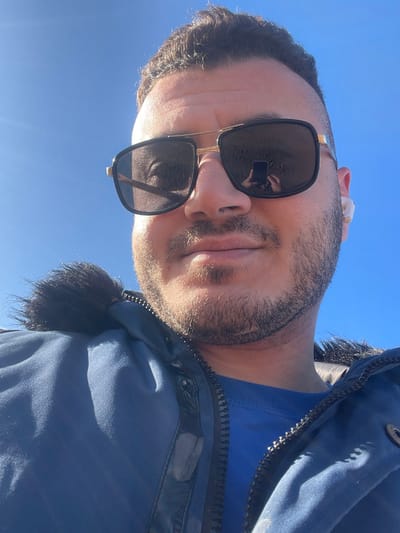 The image depicts a close-up, upward-angled outdoor self-portrait of an adult male beneath a clear, bright blue sky. The male has dark hair, a short beard, and is wearing large, dark-lensed sunglasses with gold-colored frames. A reflection of a smartphone held by a hand is visible in the left sunglass lens, indicating the subject is taking the photograph. He is dressed in a blue jacket with a dark faux fur trim on the hood, over a blue t-shirt. A white wireless earbud is present in his right ear. Strong sunlight illuminates the scene. The image provides no visual indicators to identify the location as Zarautz, Spain.