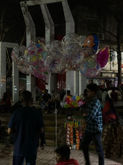 A nighttime scene captured in Narayanganj, Bangladesh, depicts a public area active with individuals and vendors. Prominently featured in the upper-center is a large cluster of clear, spherical balloons internally decorated with colorful confetti, heart shapes, and cartoon characters, including Hello Kitty. Within this cluster, opaque character balloons, such as one with a blue-haired, orange-faced figure, are also visible. These balloons are tethered by multiple thin strings extending downwards.

In the foreground, an adult male wearing a dark blue t-shirt and dark trousers stands with his back to the viewer, facing into the scene. To his right, a second adult male, partially blurred from motion, walks leftward, clad in a blue plaid shirt and dark trousers. A young individual in a red t-shirt is seated on the ground in the lower-center foreground, looking left. Near the ground, to the right of the seated individual, a display rack exhibits multiple rows of packaged snack items. Further into the mid-ground, a vendor holds a stick featuring a collection of small, multicolored balloons.

Multiple individuals are seated or resting on a series of concrete steps that lead up towards a background structure. This structure is characterized by distinct white-tiled vertical pillars or slabs. Artificial lighting illuminates parts of the scene, creating areas of brightness and deep shadow against the dark night sky. Tree branches are discernible against the sky on the upper right, and a wall with faint imagery is visible on the far right.
