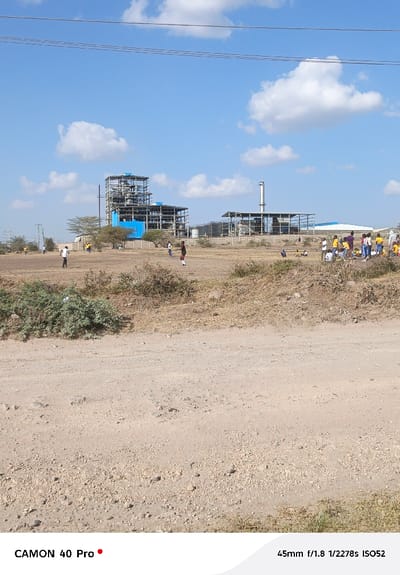 The image depicts an expansive outdoor scene featuring an industrial complex, open fields, and groups of people under a clear sky.
In the background, a large industrial facility is visible, characterized by multiple steel-framed structures. The most prominent structure on the left has complex piping and machinery within its framework, with some lower sections clad in a bright blue material. To its right, another structure also features a steel frame and a tall, slender, silver-colored smokestack or chimney rising from it. Behind these main structures, additional industrial components like storage tanks and smaller buildings can be discerned. The entire complex appears to be either under construction or recently built. A fence delineates the boundary of the industrial area.
The midground consists of a large, open, dry, and somewhat barren field. The ground is largely unpaved and dusty, with patches of dry grass and scattered, scrubby bushes and small trees. Several groups of people are dispersed across this field. On the far left, a few individuals are standing near the edge of the field. Closer to the center, two individuals, one in a white shirt and another in a black shirt, are engaged in an activity, possibly playing a game. To the right, a larger gathering of people, many dressed in bright yellow shirts or tops, are clustered together. They appear to be sitting or standing in a group, with one person near them wearing a purple top. These groups seem to be children or young adults.
In the immediate foreground, a wide, unpaved dirt road or pathway stretches across the bottom of the frame. It is composed of light-colored soil and small rocks. Some sparse, dry vegetation grows along its edges.
The sky is a bright, clear blue with scattered, fluffy white cumulus clouds, indicating a sunny day. High above, several thin, dark lines, likely power lines, stretch horizontally across the upper portion of the sky. The overall lighting suggests it is late morning or early afternoon.
Text visible in the bottom left corner reads "CAMON 40 Pro," indicating the camera model used. In the bottom right corner, the camera settings "45mm f/1.8 1/2278s ISO52" are displayed.