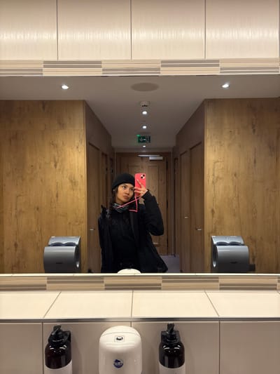 A female subject is depicted taking a mirror selfie in a modern public washroom located in St. Christoph, Austria. The subject, positioned centrally, has dark, curly hair visible under a black beanie and wears a black jacket over a dark top, with pink earbuds draped around her neck. She holds a bright pink-cased smartphone in her right hand, pointed towards the mirror to capture her reflection. The washroom features light-colored wood paneling on the walls, a white countertop below the mirror, and white tiled walls underneath the counter, accented with horizontal grey and beige stripes. Three soap dispensers are visible on the tiled wall: two dark brown pump bottles flanking a central white dispenser. Two grey Dyson-style hand dryers are mounted on the wood-paneled walls, one on each side of the subject's reflection. The reflection in the mirror reveals a brightly lit hallway or corridor with additional wood-paneled doors and overhead recessed lighting, including a green emergency exit sign in the distance. The overall environment suggests a well-maintained facility.