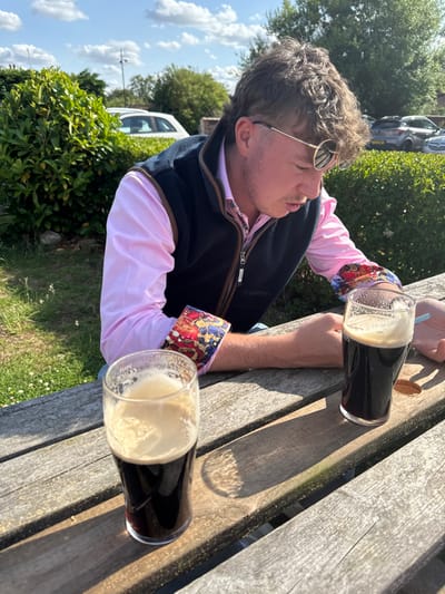 A man with curly hair and sunglasses sits at a wooden table with two glasses of dark beer. He wears a pink shirt with patterned cuffs and a navy vest. The scene is outdoors on a sunny day, with trees and parked cars in the background. The table's shadow and the beer glasses' shadows are visible.