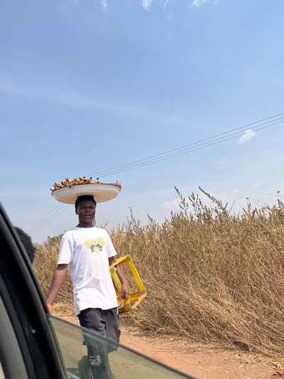 The image depicts a young adult male street vendor in Nandu, Nigeria, captured from the perspective of a vehicle. He stands on what appears to be a dirt road shoulder, positioned between the vehicle and a backdrop of dry, tall golden-brown vegetation. The man has a dark complexion and short, dark hair. He wears a white short-sleeved t-shirt with a gold-colored "GUCCI" logo featuring two stylized animal heads forming a heart shape, and dark-colored shorts or trousers. A large, circular white tray, filled with numerous small, golden-brown fried food items (likely snacks), is balanced on his head. In his left hand, he carries a bright yellow, empty plastic crate or frame. His mouth is slightly open, and he is looking directly towards the camera. The sky above is clear and bright blue with a few faint white clouds, and utility lines are visible crossing the upper background. The overall environment suggests a sunny, dry daytime setting.
