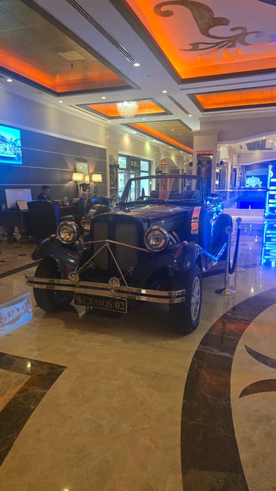 A vintage, dark blue convertible automobile is displayed indoors on a polished marble floor. The vehicle is positioned in the foreground, facing slightly to the right of the camera. It features prominent chrome accents, including the headlights, bumper, and trim. A license plate on the front bumper reads "CRATOS 02". Behind the car, the environment appears to be a luxurious hotel lobby. The ceiling is elaborately decorated with recessed lighting and gold accents, casting an orange glow. A chandelier hangs from the ceiling, and modern furniture, including desks and chairs with people seated at them, are visible in the background. The overall impression is one of opulence and a blend of classic and modern design. The location is specified as Kazafani, Northern Cyprus / Cyprus, suggesting this is likely a decorative display within a hotel or entertainment venue in that area.