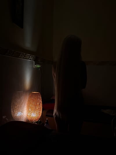 In a dimly lit room, possibly a bedroom or spa, in Khan Boeng Keng Kang, Cambodia, a person with long blonde hair stands with their back to the viewer. They are silhouetted against a wall, and their body appears nude. The room is furnished with two woven lamps that emit a warm, orange glow, casting intricate patterns of light and shadow on the wall and floor. One lamp is positioned to the left of the standing figure, while the other is closer to the foreground. A framed picture hangs on the wall in the upper left corner, partially obscured by shadow. A green object, possibly an air freshener or night light, is plugged into a wall socket. The overall atmosphere is intimate and serene.