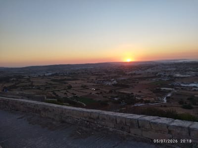 A sunset is captured over the town of Għargħur, Malta. The sky transitions from a light blue at the top to shades of orange and yellow near the horizon. The sun is a bright orb low on the horizon, casting a warm glow. In the foreground, a low stone wall made of rough-hewn blocks runs horizontally across the frame. The ground in front of the wall is a paved surface with some debris scattered on it. The middle ground and background reveal a landscape of fields, some with cultivated patches, interspersed with scattered buildings and trees. The overall impression is of a rural setting viewed from an elevated position. A timestamp "05/07/2026 20:18" is visible in the bottom right corner.