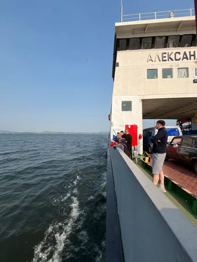 The image captures a view from the deck of a ferry traveling on a body of water, likely the Krasnoyarsk Reservoir, near the village of Novoselovo, Russia. The ferry has the Cyrillic name "АЛЕКСАНДР" visible on its side. Several people are standing on the ferry's deck, some facing the water and others conversing. A dark-colored car is parked on the ferry's deck, along with what appears to be a white van or bus. The water is choppy, indicating the ferry is in motion, and a wake is visible trailing behind. The sky is clear and blue, with a distant shoreline featuring hills and some buildings, consistent with the landscape around Novoselovo. The overall scene suggests a mode of transportation and leisure activity common in the region.