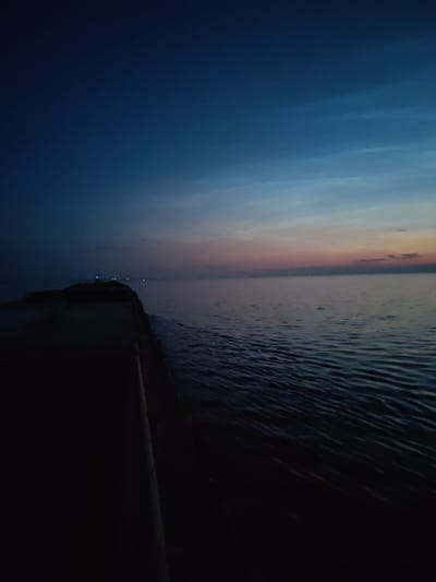 The image depicts a pier extending into a body of water at dusk. The sky is a deep blue, transitioning to lighter shades with hints of pink and orange near the horizon, indicating the last moments of daylight. The water is dark and slightly rippled. Lights are visible in the distance along the horizon, suggesting a coastline or other structures. The pier itself is dark and appears to be made of concrete. There are no people visible in the image. The scene conveys a sense of solitude and the transition from day to night. The location is specified as Pochinok, Russia.